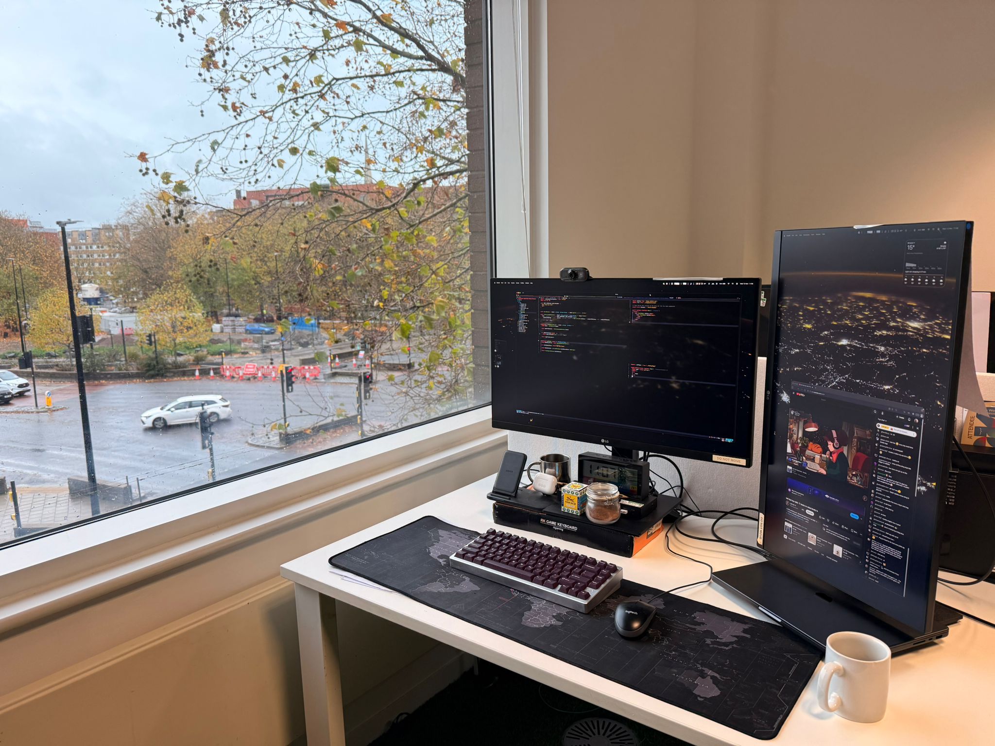 Picture of two screens, a keyboard and mouse on a white desk, with a rainy view of of a window